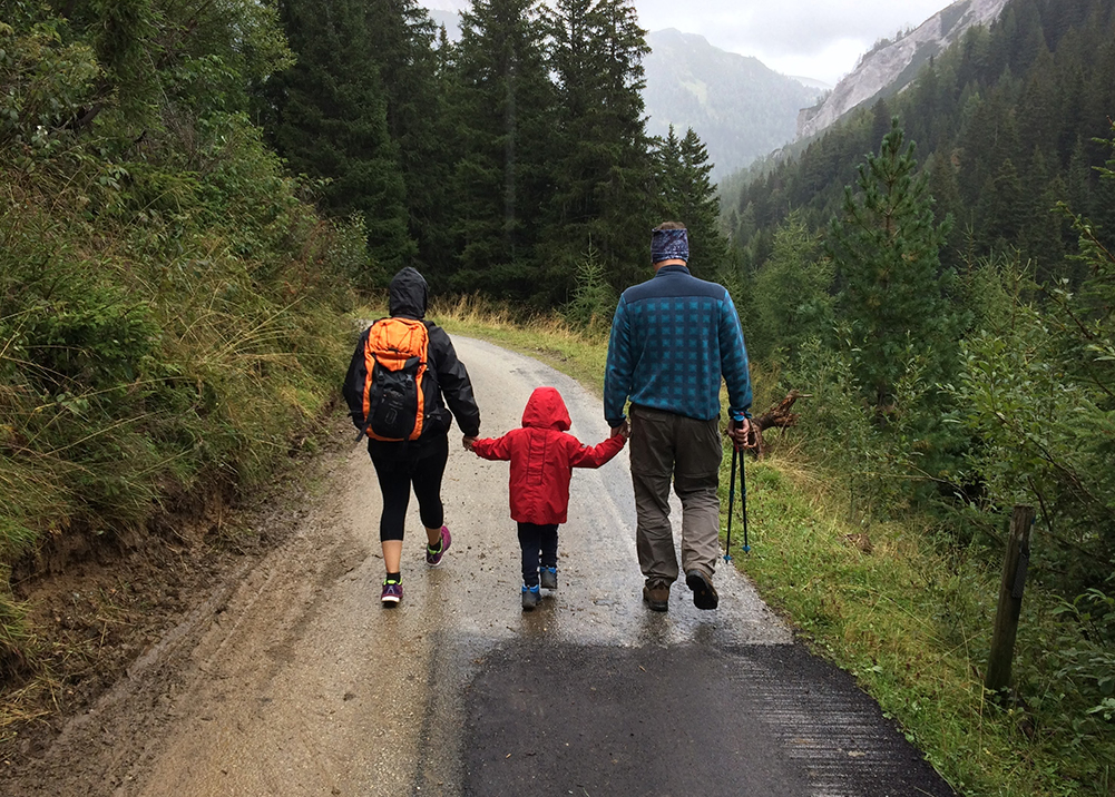 A family of three is hiking on a wet path. They are wearing jackets, active clothing and hiking shoes.