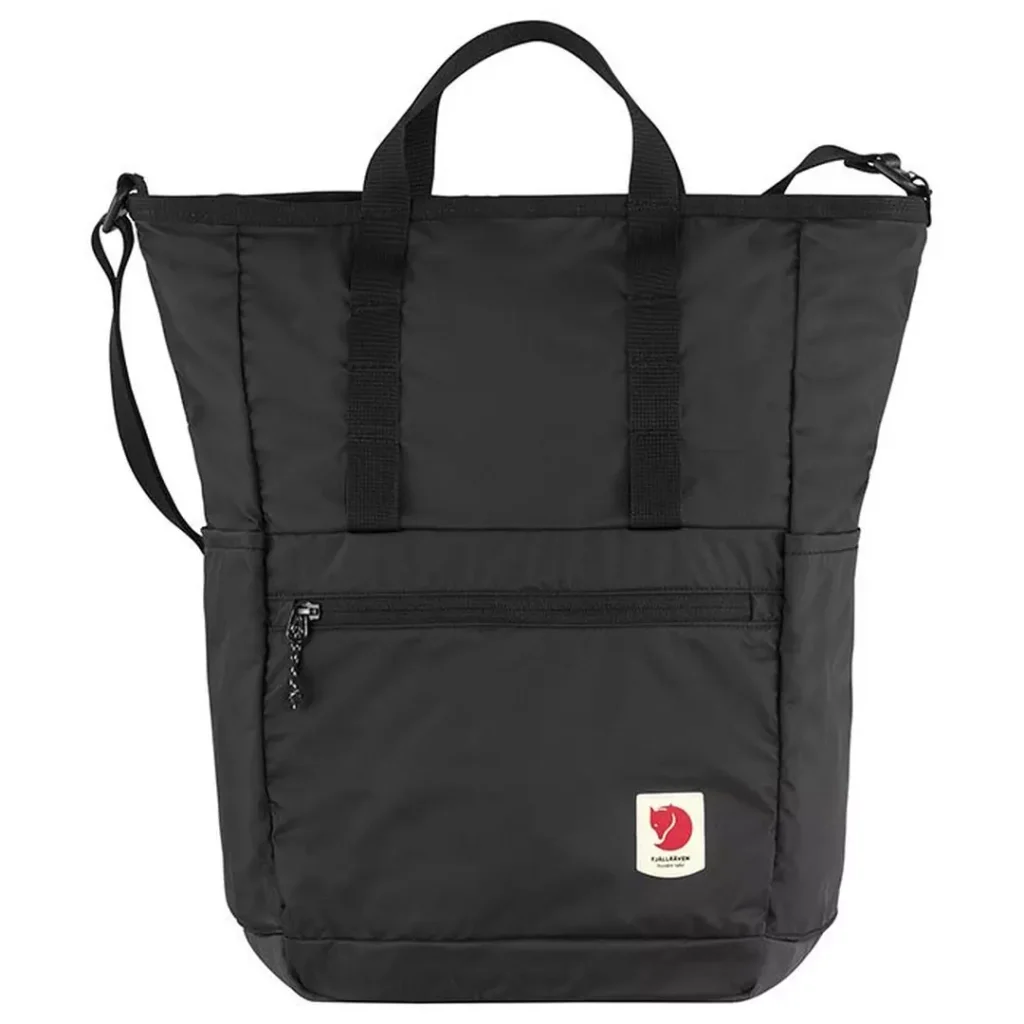 black rectangular Fjallraven tote/backpack with black webbing carry handles and a shoulder strap, front zip accessories pocket and a small contrast white/red Fjallraven fox logo patch at the bottom-right