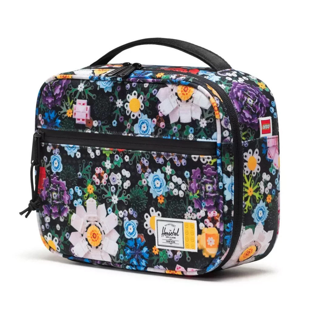 colourful, rectangular Herschel lunch bag with a limited-edition LEGO floral pattern, a top carry handle and a front zip pocket, plus a Herschel x LEGO label at the bottom-right side