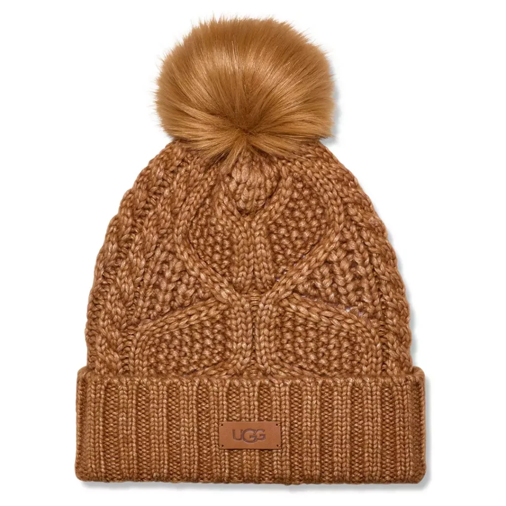 Chestnut brown toque-style UGG winter hat with a unique cable knit pattern and a ribbed cuff with a small tonal brown rectangular leather logo patch at the cuff and a soft, fluffy faux fur pompom at the top