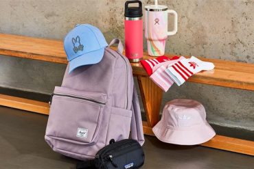a wooden bench with a light purple/greyish Herschel backpack in front, a blue Psycho Bunny baseball cat on top of the backpack, a light pink adidas bucket hat below the bench, to the right of the backpack; a pink YETI tumbler on top of the bench next to a light pink Stanley straw tumbler and 3 different adidas crew socks