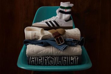 dark teal plastic stadium-style chair against a dark wood background with a folded pile of clothing with a Herschel sweatshirt at the bottom, a diamond-quilted jacket above, a folded pair of jeans above that, a cable knit sweater folded above the jeans with a belt and an adidas Samba shoe on top and striped Polo crew socks tucked into the sneaker