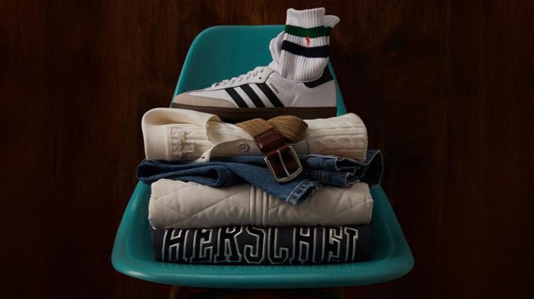 dark teal plastic stadium-style chair against a dark wood background with a folded pile of clothing with a Herschel sweatshirt at the bottom, a diamond-quilted jacket above, a folded pair of jeans above that, a cable knit sweater folded above the jeans with a belt and an adidas Samba shoe on top and striped Polo crew socks tucked into the sneaker