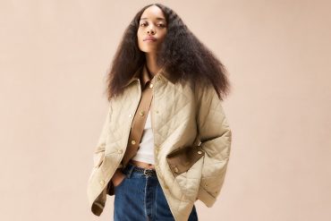 female model in the centre of the frame against a neutral-coloured tan background; she is wearing dark blue jeans with a white top layered under a tan lightweight jacket with diamon quilting allover and contrast brown pocket flaps at the lower-front body