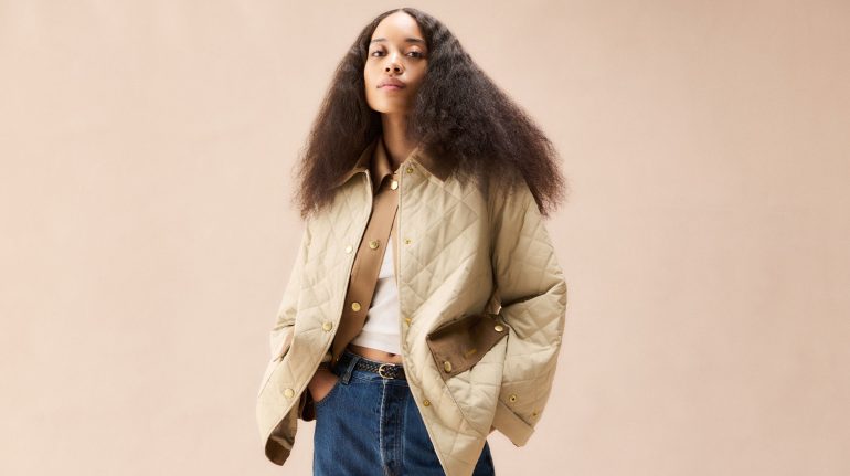 female model in the centre of the frame against a neutral-coloured tan background; she is wearing dark blue jeans with a white top layered under a tan lightweight jacket with diamon quilting allover and contrast brown pocket flaps at the lower-front body