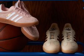 two pairs of adidas sneakers in muted pastel colours (light pink and tan/beige) sit on a blue metal ball rack with a classic brown basketball to the left and a dark wood wall in the background