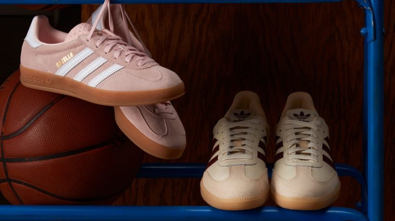 two pairs of adidas sneakers in muted pastel colours (light pink and tan/beige) sit on a blue metal ball rack with a classic brown basketball to the left and a dark wood wall in the background